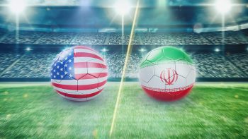 USA vs Iran World Cup country flags portrayed on two soccer balls side by side on an empty soccer field with three strobe lights pointing at them